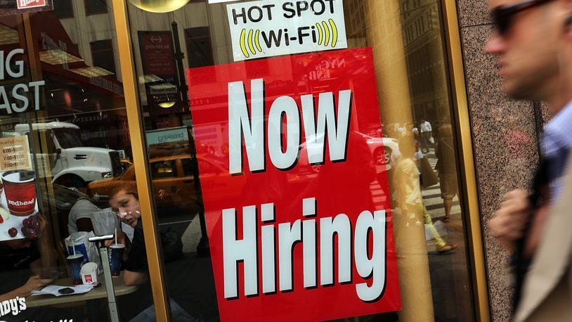 A "now hiring" sign is viewed in the window of a business.