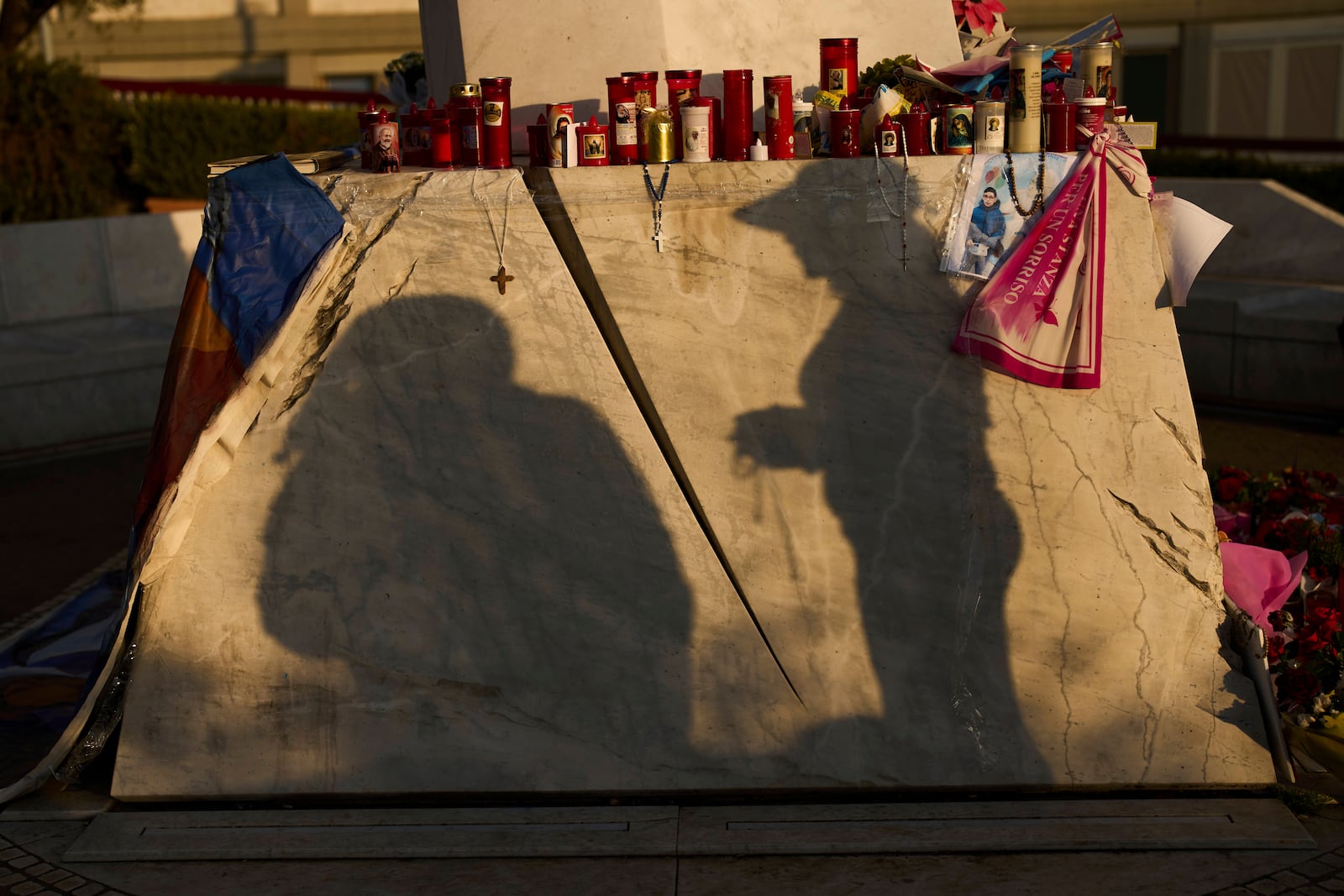 A woman is silhouetted as she holds a Christian rosary after praying next to the statue of Pope John Paul II placed outside the Agostino Gemelli hospital in Rome, Thursday, March 6, 2025. (AP Photo/Francisco Seco)