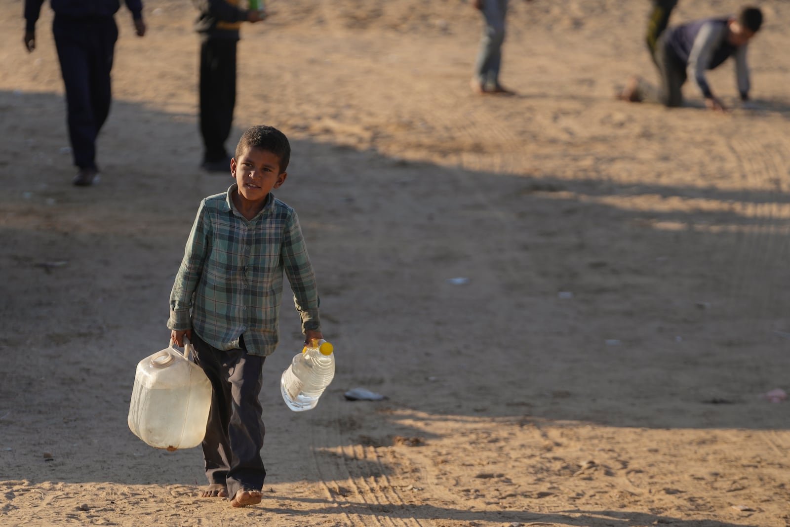 A Palestinian boy carries jerrycans of water along a dirt road in Deir al-Balah, Gaza Strip, Saturday Dec 21, 2024 (AP Photo/Abdel Kareem Hana)