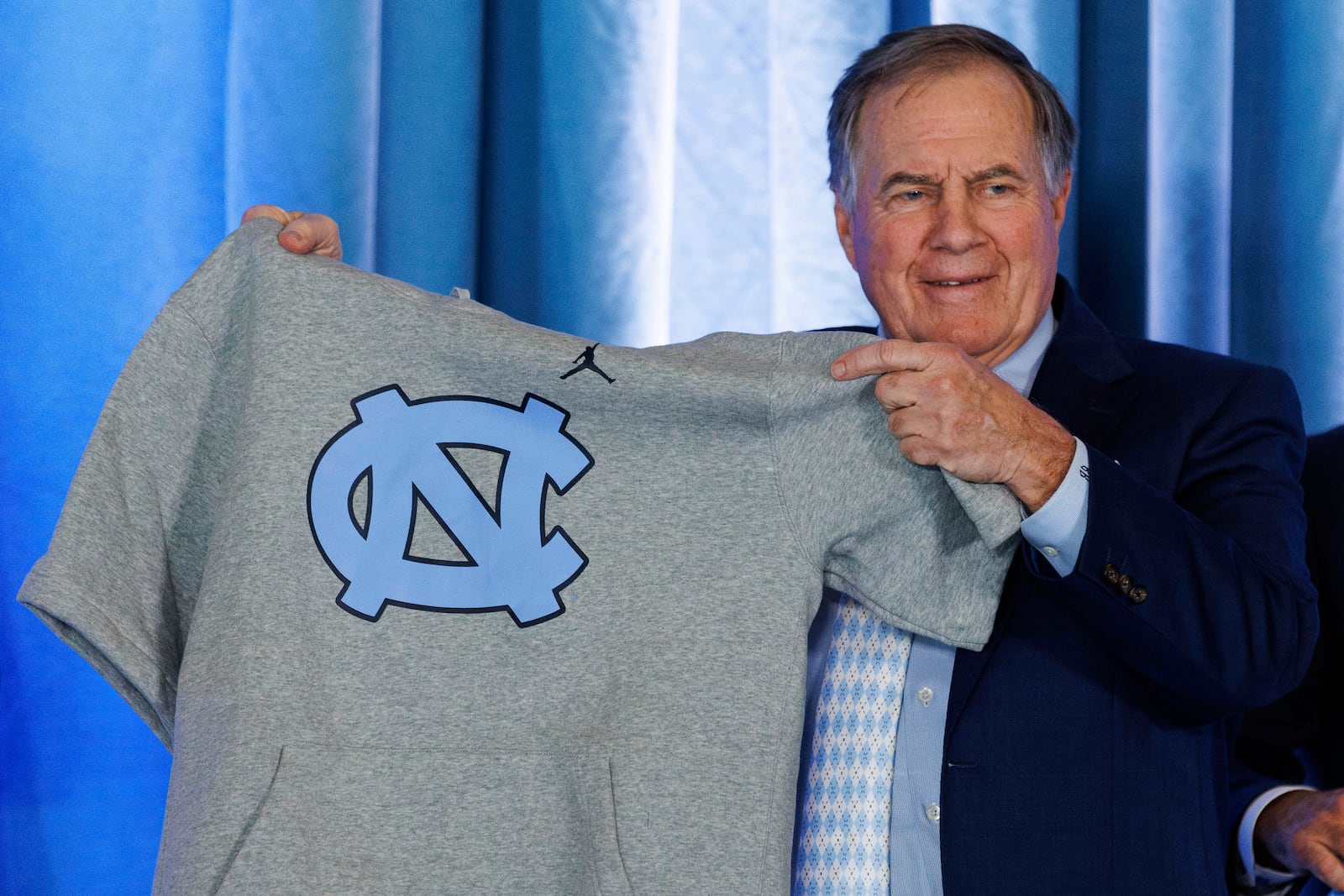 New North Carolina football coach Bill Belichick holds up a UNC branded sleeveless hoodie presented to him during an NCAA college football press conference announcing his hiring, Thursday, Dec. 12, 2024, in Chapel Hill, N.C. (AP Photo/Ben McKeown)