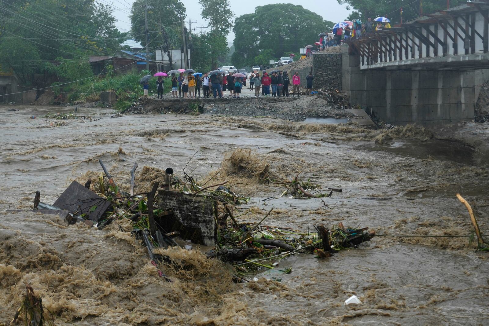 Residents stand alongside the banks of a river overrun by rains brought on by Tropical Storm Sara, in the Flor de Cuba neighborhood, on the outskirts of San Pedro Sula, Honduras, Saturday, Nov. 16, 2024. (AP Photo/Moises Castillo)