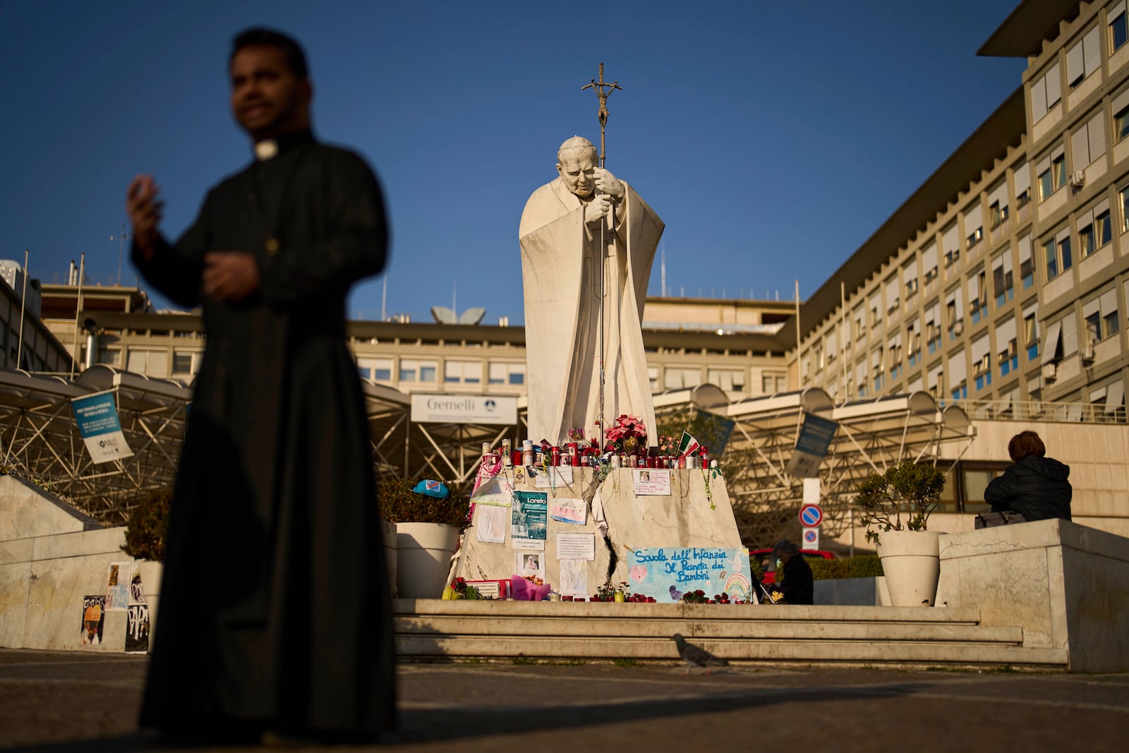 People and a Christian priest stand next to the statue of Pope John Paul II outside the Agostino Gemelli hospital in Rome, Thursday, March 6, 2025. (AP Photo/Francisco Seco)