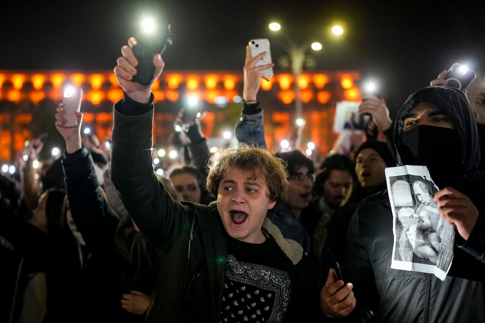 Youngsters shout slogans and flash the light of their mobile phones in Bucharest, Romania, Wednesday, Nov. 27, 2024, next to an altered version of a classic painting, depicting Russian President Vladimir Putin and Calin Georgescu, the independent candidate for Romanian presidency who won the first round of elections making it to the Dec. 8, runoff. (AP Photo/Vadim Ghirda)