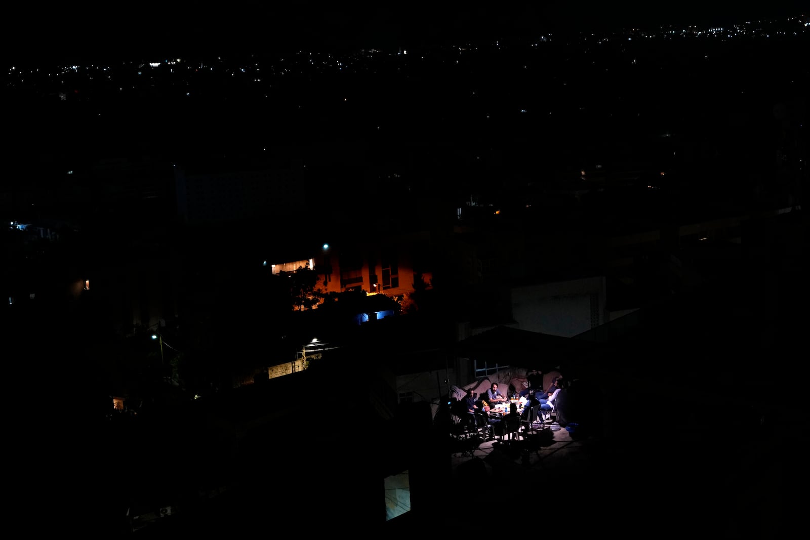 Residents set on the roof of a building and have dinner as Dahiyeh suburb, background, remains in darkness after Israeli airstrikes, Lebanon, Friday, Oct. 11, 2024. (AP Photo/Hassan Ammar)