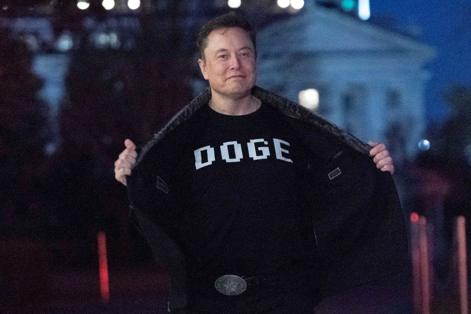 Elon Musk flashes his t-shirt that reads "DOGE" to the media as he walks on South Lawn of the White House, in Washington, Sunday, March 9, 2025. (AP Photo/Jose Luis Magana)