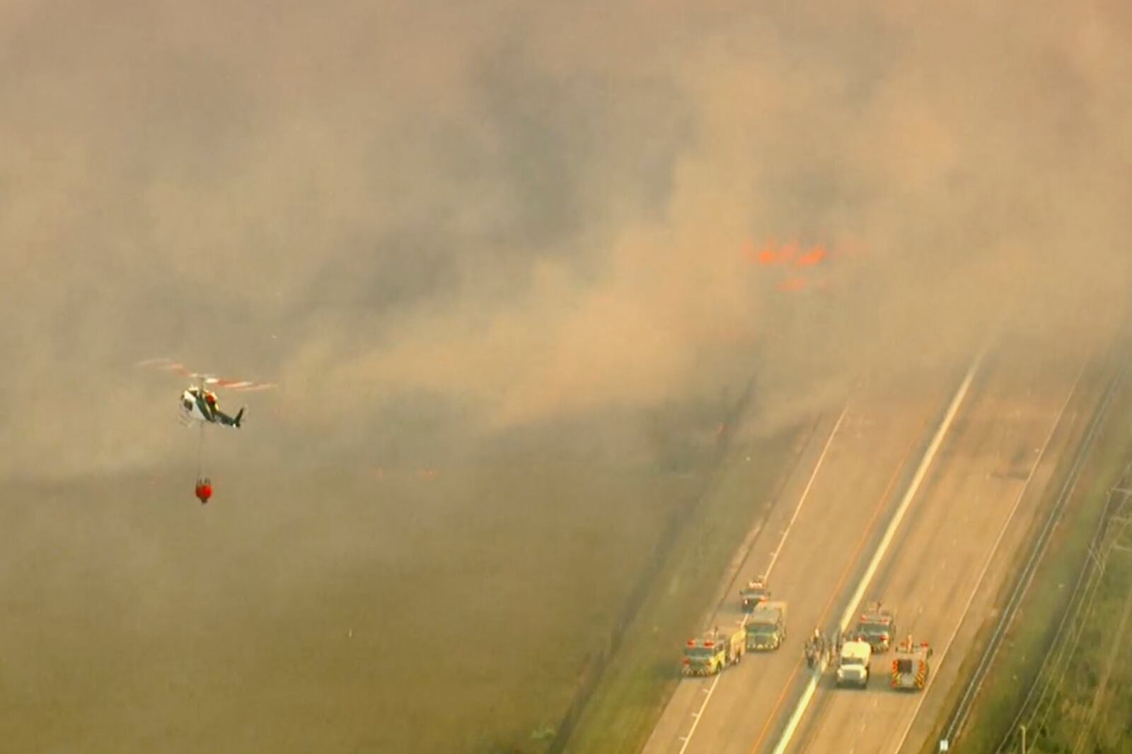 In this image taken from video provided by WSVN-TV, a brush fire burns along U.S. 1 as a helicopter carrying a water tank flies toward the fire Tuesday, March 18, 2025, south of Dade County, Fla. (WSVN-TV via AP)