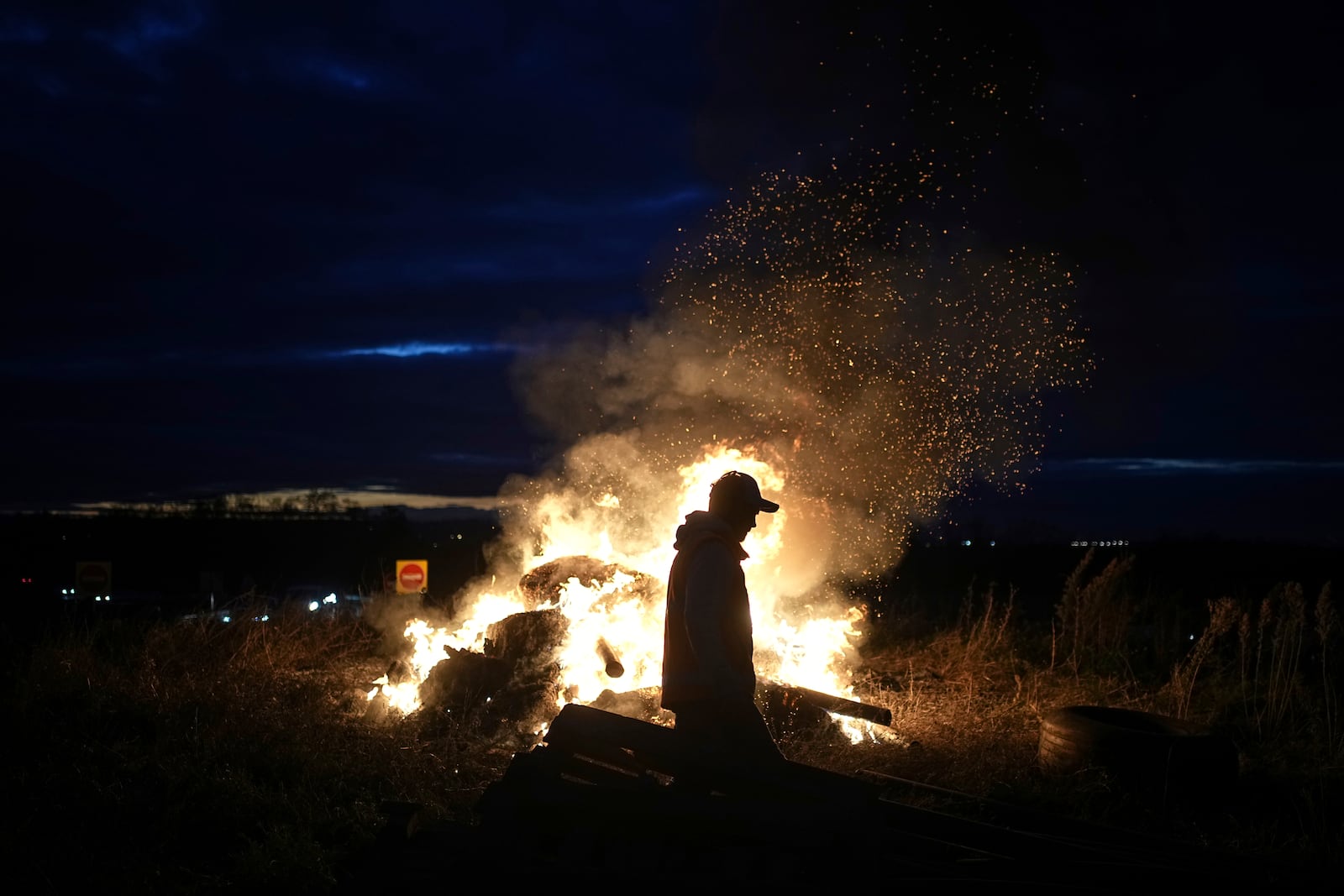 A farmer walks by a fire as farmers protest against the EU-Mercosur trade agreement, in Saint-Laurent-de-Mure, near Lyon, central France, Monday, Nov. 18, 2024. (AP Photo/Laurent Cipriani)