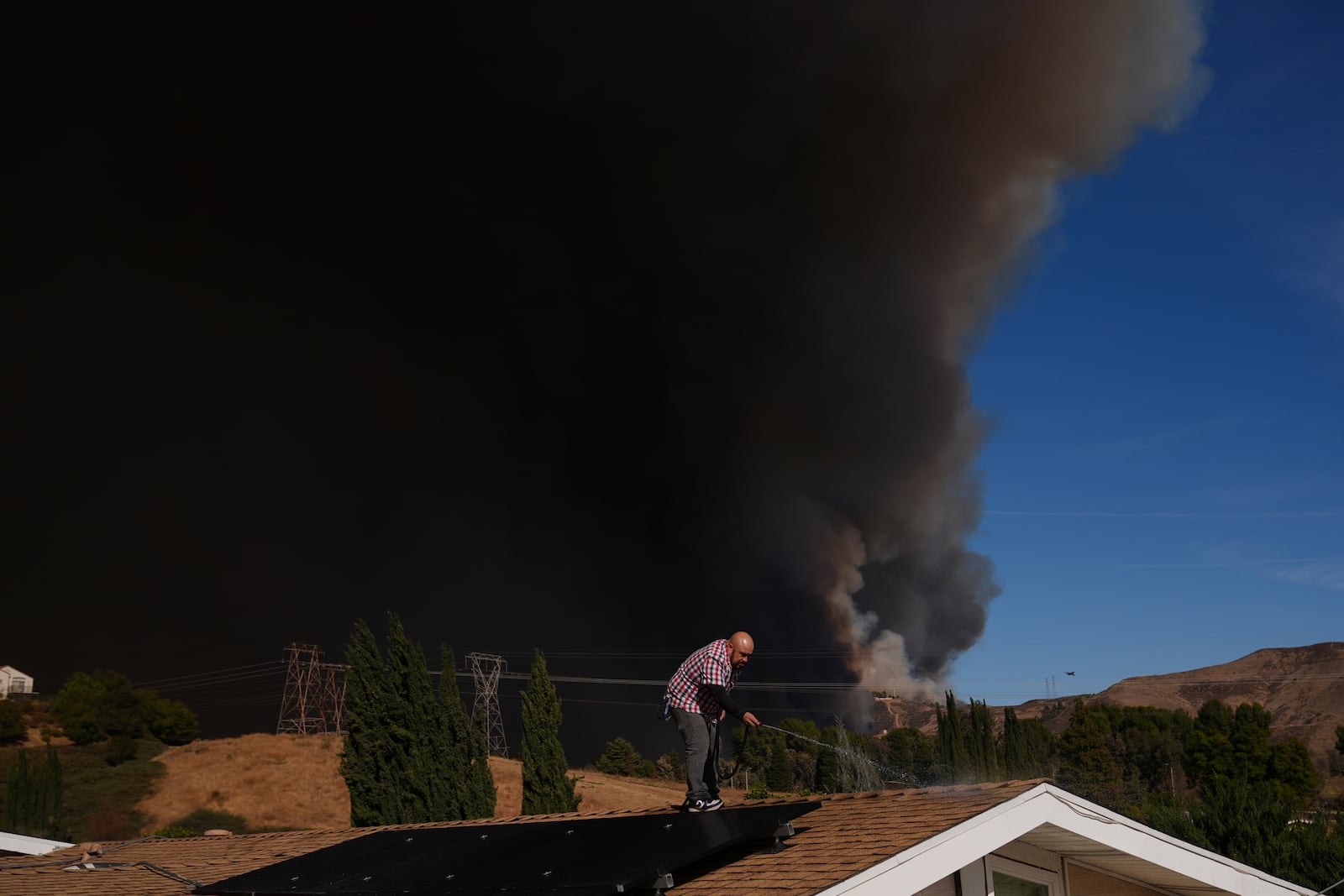 A home owner sprays water from the top of the roof at his brother's home Castaic, Calif., as a large plume of smoke caused by the Hughes Fire rises from Castaic Lake Wednesday, Jan. 22, 2025. (AP Photo/Marcio Jose Sanchez)