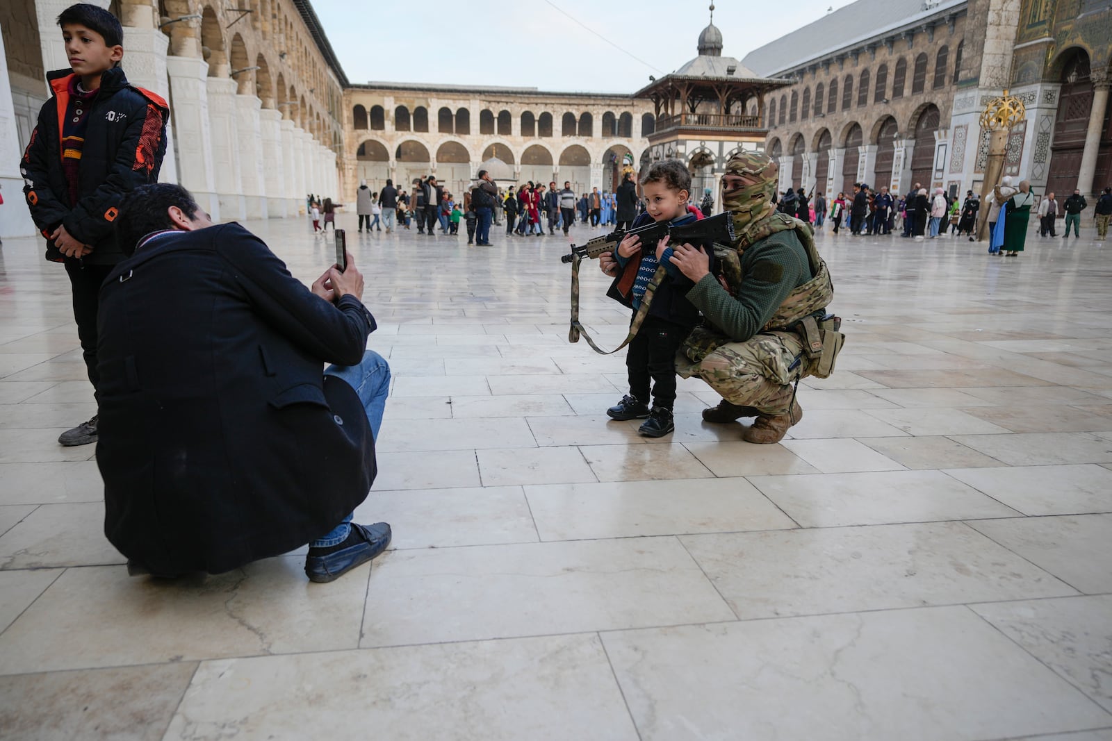 A boy poses for a photo with a masked Hayat Tahrir al-Sham (HTS) fighter holding a gun in the courtyard of the Umayyad Mosque in the old walled city of Damascus, Syria, Tuesday, Dec. 10, 2024. (AP Photo/Hussein Malla)