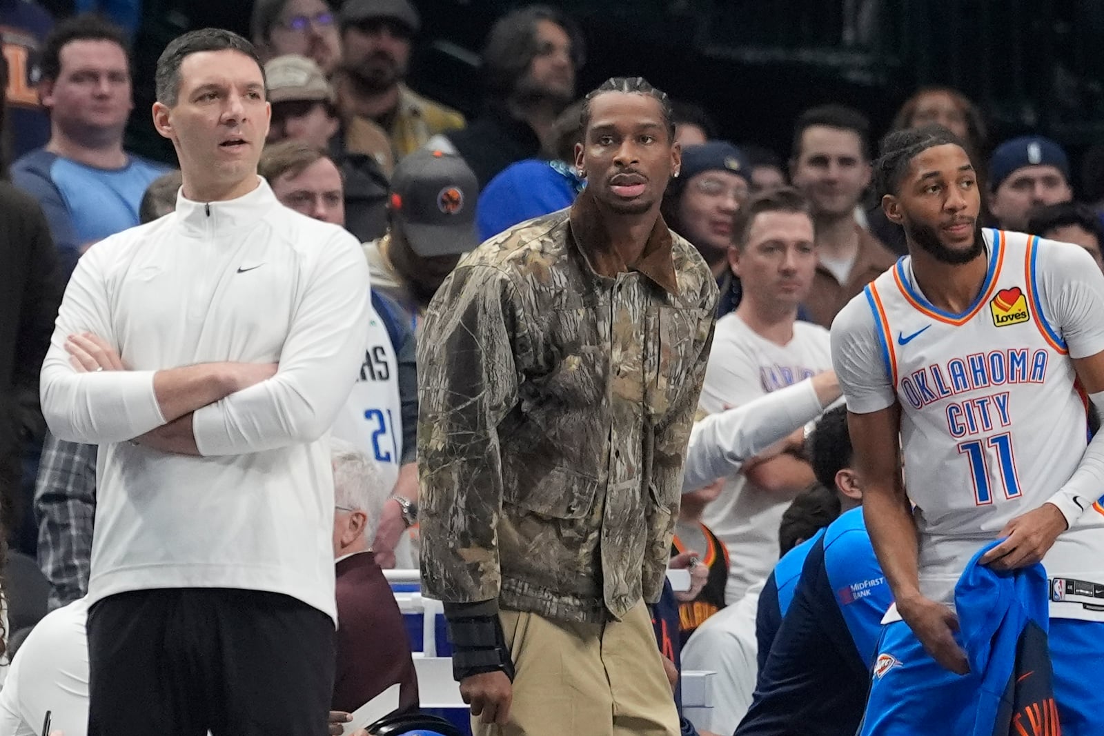 Injured Oklahoma City Thunder guard Shai Gilgeous-Alexander, center, looks on from the sideline with head coach Mark Daigneault, left, and teammate guard Isaiah Joe (11) during the first half of an NBA basketball game against the Dallas Mavericks, Friday, Jan. 17, 2025, in Dallas. (AP Photo/LM Otero)