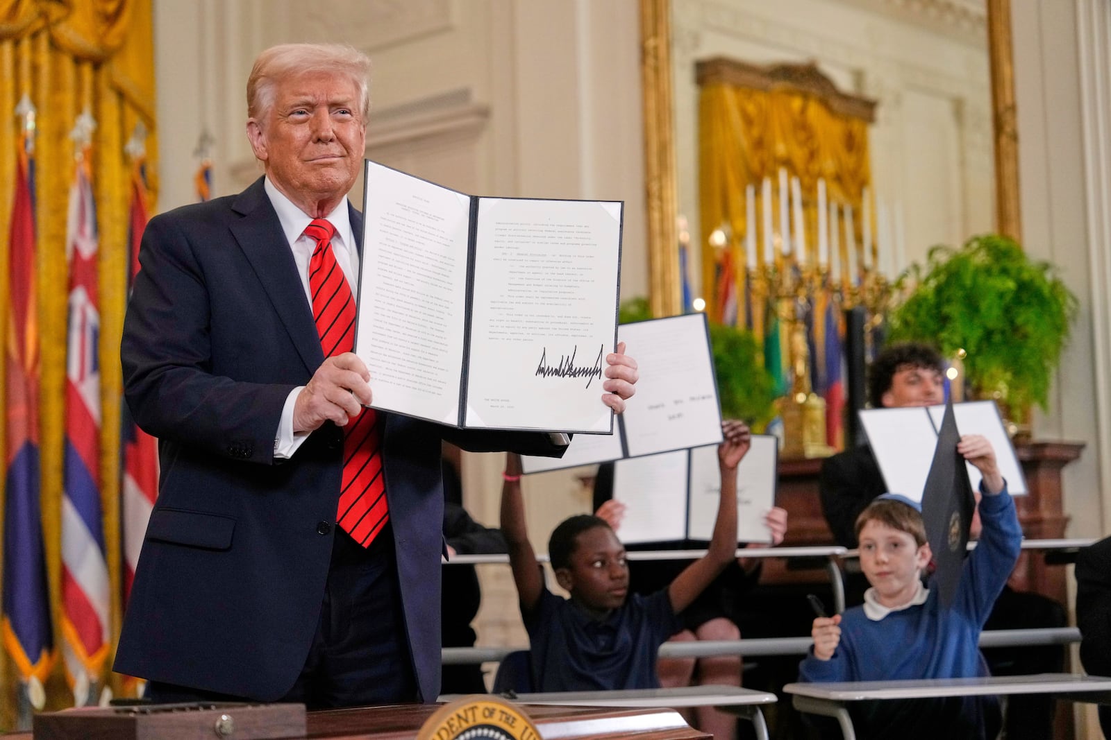 President Donald Trump, left, holds up a signed executive order as young people hold up copies of the executive order they signed at an education event in the East Room of the White House in Washington, Thursday, March 20, 2025. (AP Photo/Ben Curtis)