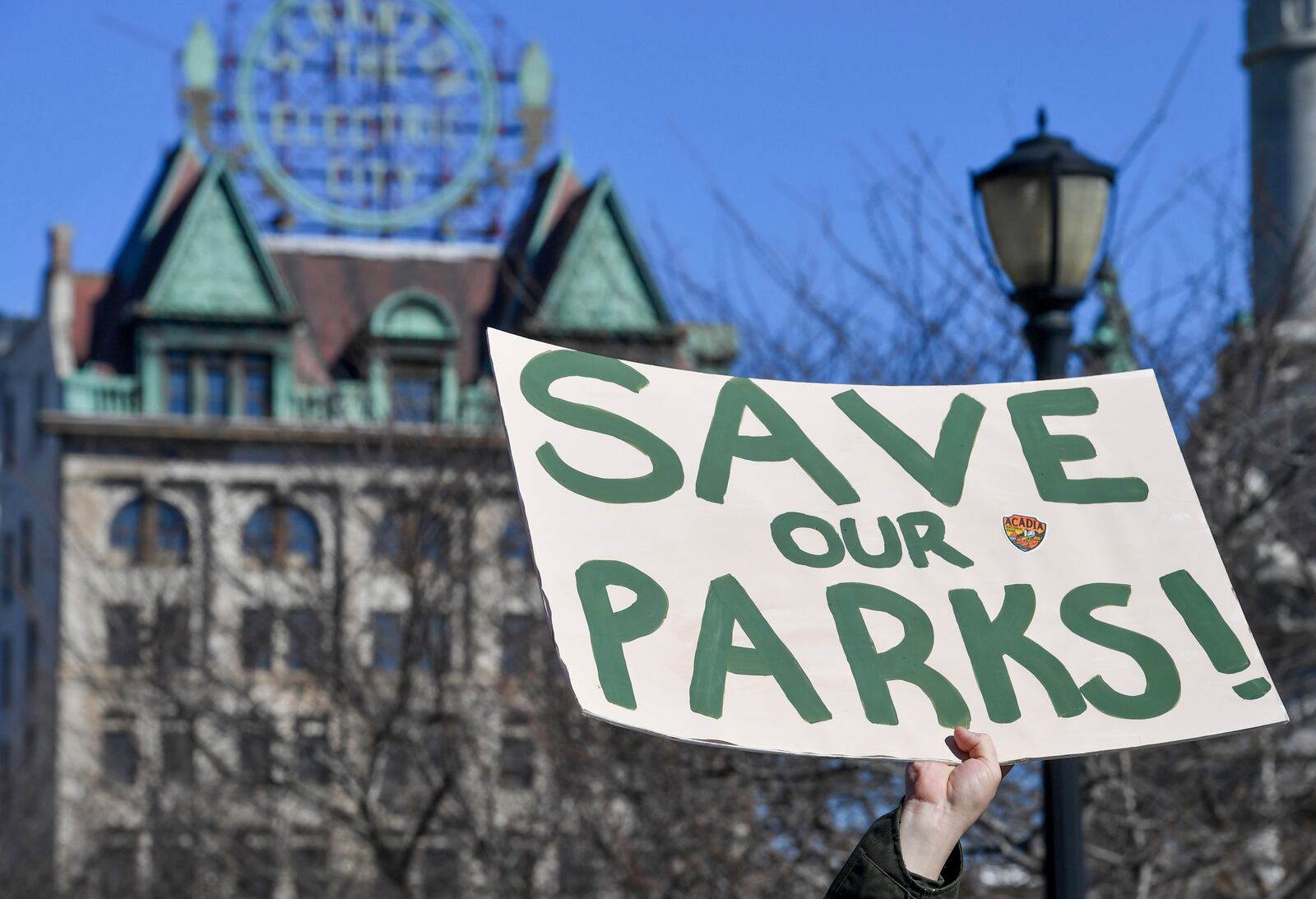 A protester raises a sign that reads "Save Our Parks" under the Scranton Electric City building at a "Save Steamtown" rally to protest the Trump administration layoffs at the Steamtown National Historic Site in Scranton, Pa., Saturday, Feb. 22, 2025. (Aimee Dilger/WVIA via AP)