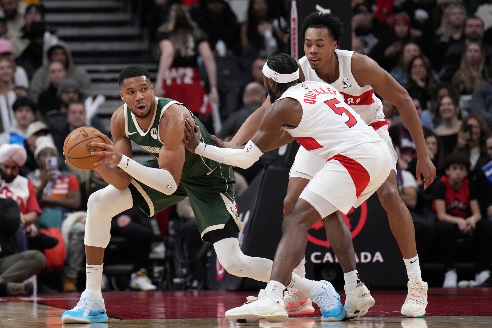 Milwaukee Bucks forward Giannis Antetokounmpo, left, looks to pass the ball while under pressure from Toronto Raptors guard Immanuel Quickley (5) and Scottie Barnes, right, during first-half NBA basketball game action in Toronto, Monday, Jan. 6, 2025. (Nathan Denette/The Canadian Press via AP)