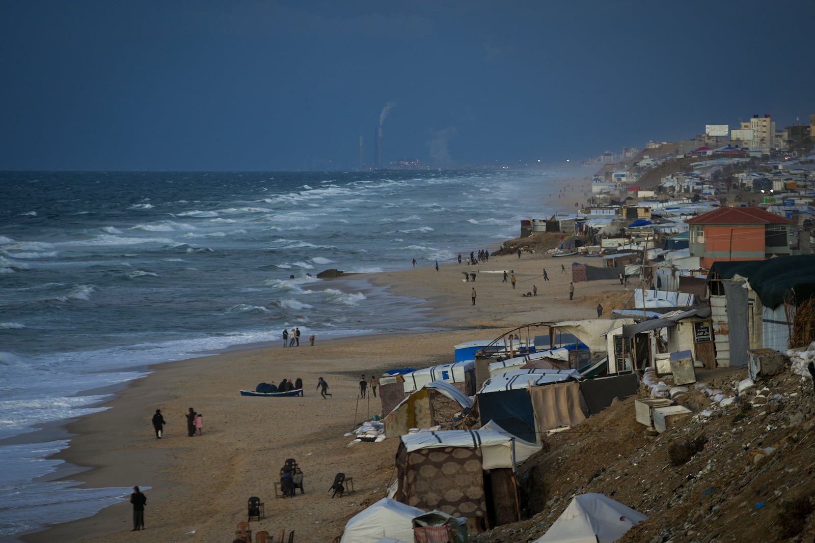 People walk along the beach next to a tent refugee camp for displaced Palestinians in Deir al-Balah, central Gaza Strip, Monday, Dec .30, 2024. (AP Photo/Abdel Kareem Hana)