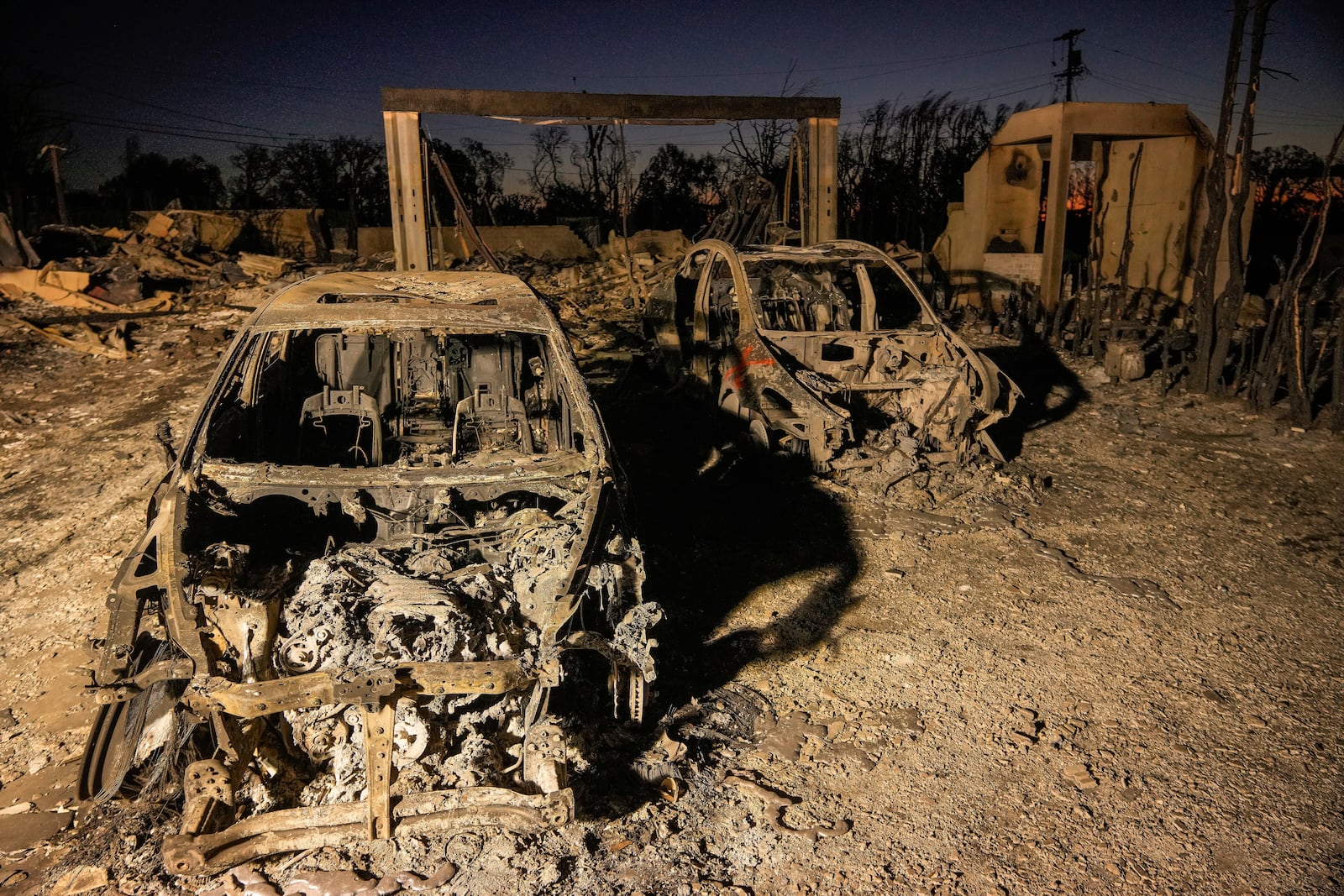 Charred vehicles are illuminated by utility lights at homes destroyed by the Palisades Fire in the Pacific Palisades neighborhood of Los Angeles, Thursday, Jan. 16, 2025. (AP Photo/Damian Dovarganes)