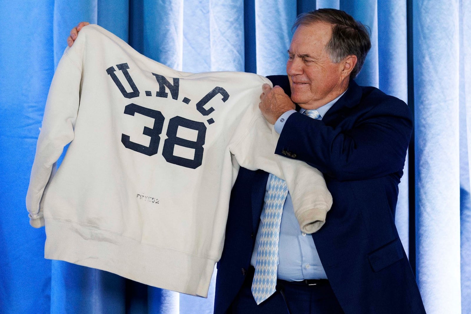 New North Carolina coach Bill Belichick holds up a sweatshirt that belonged to his father when he was on the coaching staff at UNC, Thursday, Dec. 12, 2024, during an NCAA college football press conference in Chapel Hill, N.C. (AP Photo/Ben McKeown)