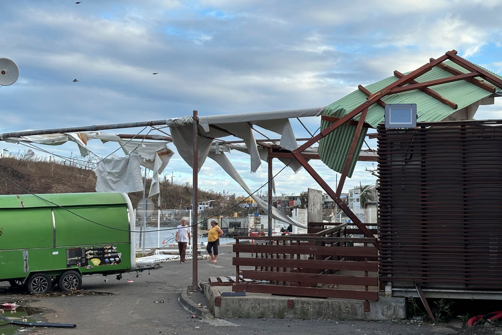 People walk by damaged constructions in Mamoudzou, in the French Indian Ocean territory of Mayotte, Monday, Dec.16, 2024 as France uses ships and military aircraft to rush rescue workers and supplies after the island group was battered by its worst cyclone in nearly a century. (AP Photo/Rainat Aliloiffa)