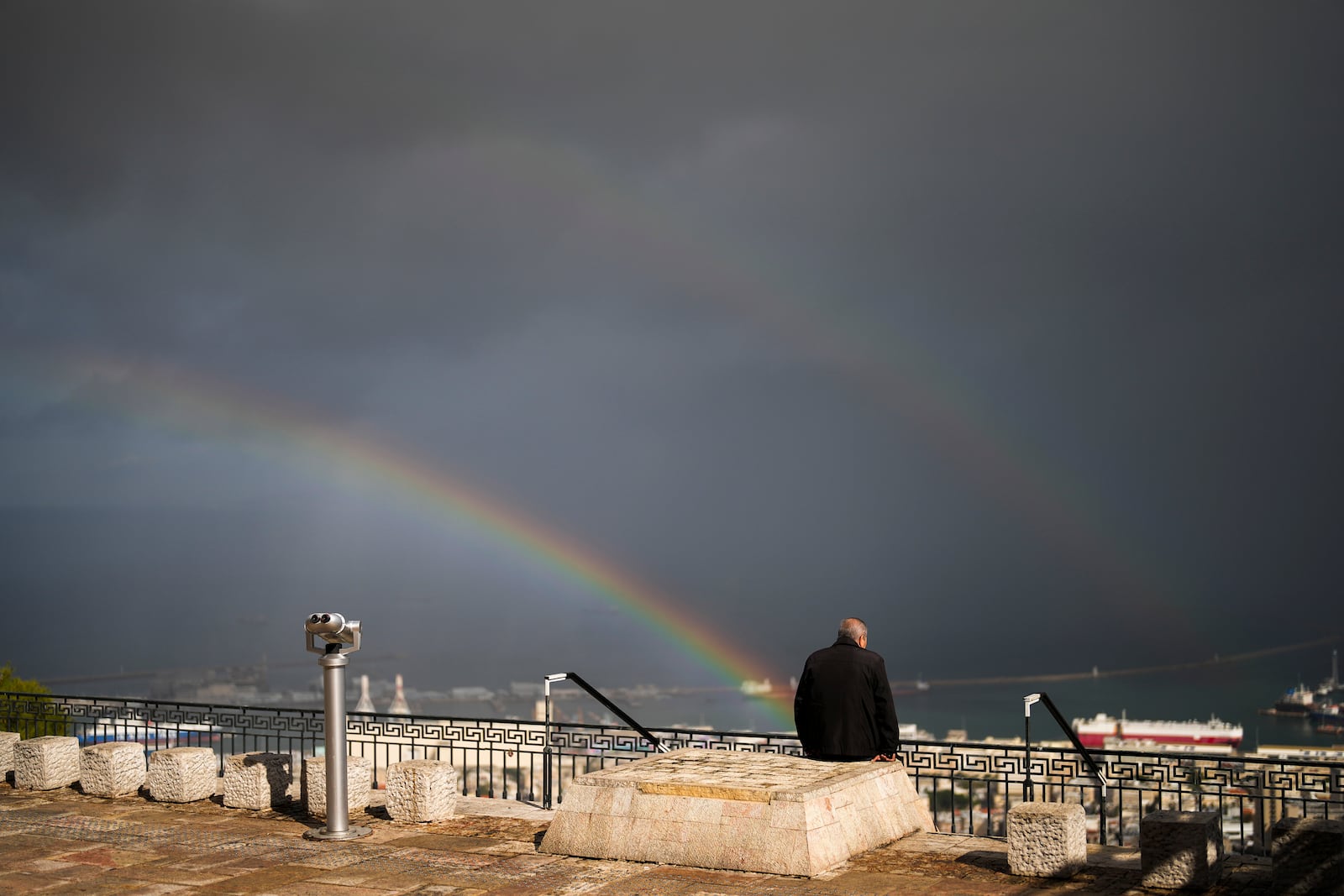 A rainbow stretches across the sky as a man sits in a promenade overlooking Haifa, Israel, Tuesday, Nov. 26, 2024. (AP Photo/Francisco Seco)