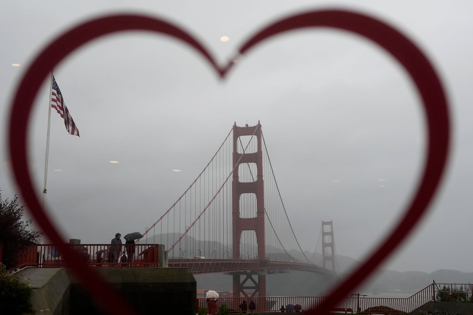 People carrying umbrellas while visiting the Golden Gate Bridge are seen through a heart on the window at the Round House Cafe in San Francisco, Wednesday, Nov. 20, 2024. (AP Photo/Jeff Chiu)