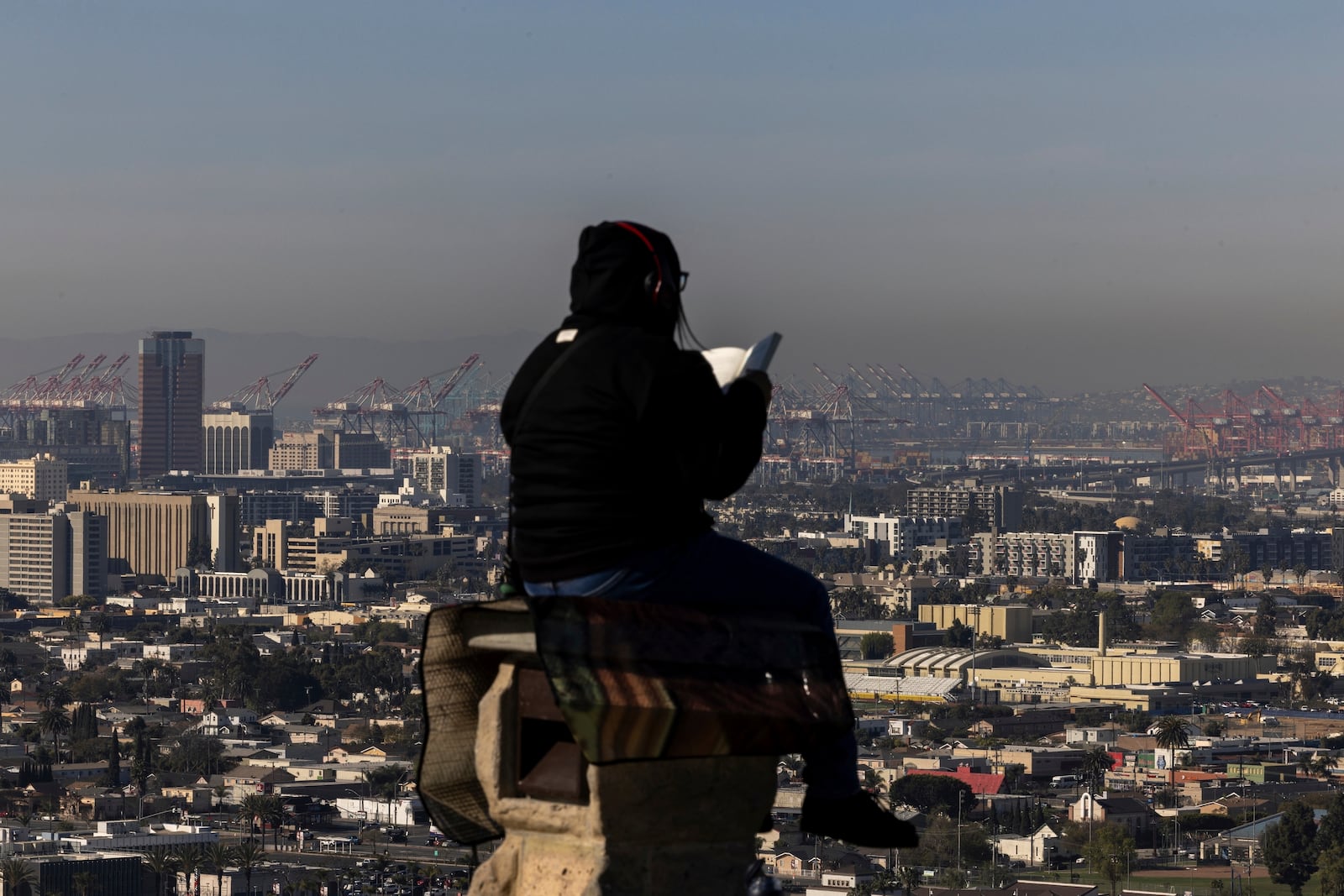 A woman reads a book sitting on a column in Hilltop Park overlooking the Ports of Long Beach and Los Angeles, Monday, March 10, 2025, in Signal Hill, Calif. (AP Photo/Etienne Laurent)