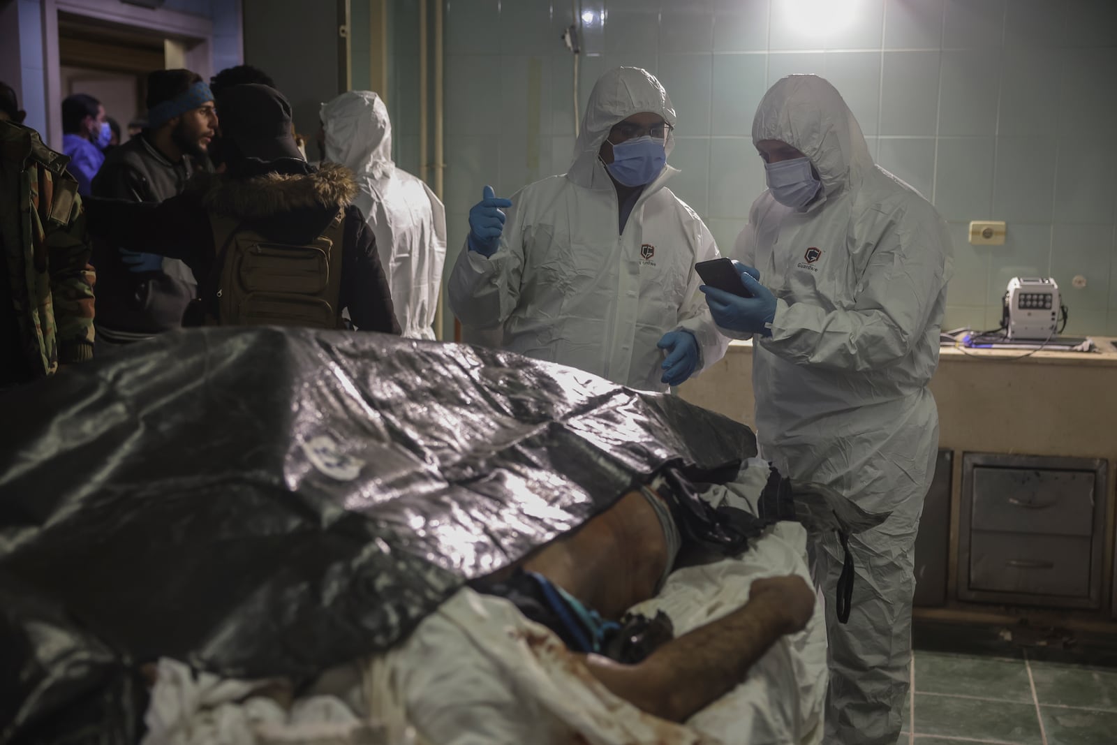 Forensic workers stand next to a body as people wait outside the morgue to identify missing relatives or friends at the Al-Mujtahid hospital in Damascus, Syria, Tuesday, Dec. 10, 2024. (AP Photo/Ghaith Alsayed)