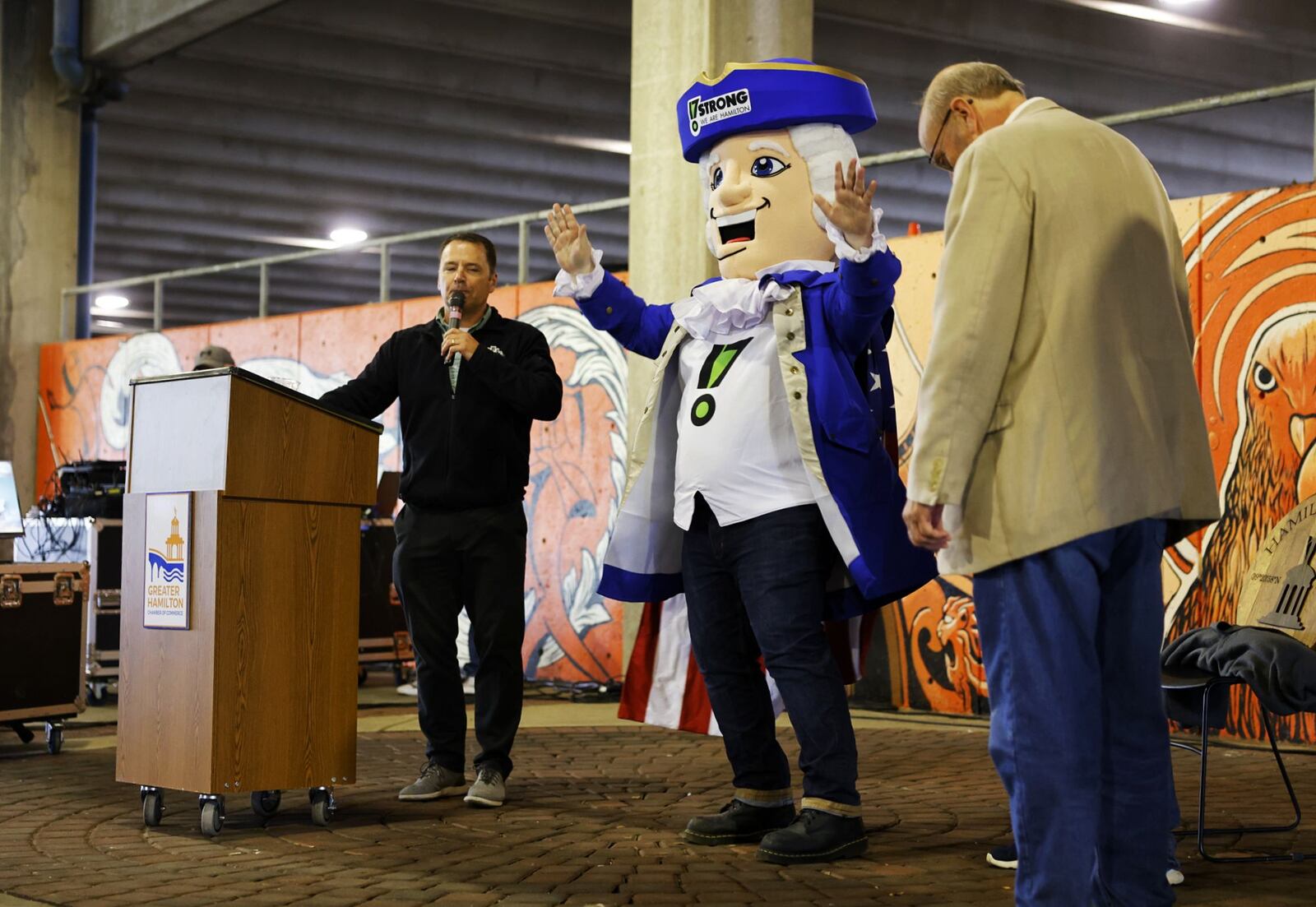 Xander, the 17 Strong mascot, stands between Jim Goodman with Municipal Brew Works, left, and Hamilton mayor Pat Moeller, right, during the state of the city address under the McDulin Parking Garage Thursday, May 5, 2022. The location was moved from the original location of Municipal Brew Works due to rain. NICK GRAHAM/STAFF