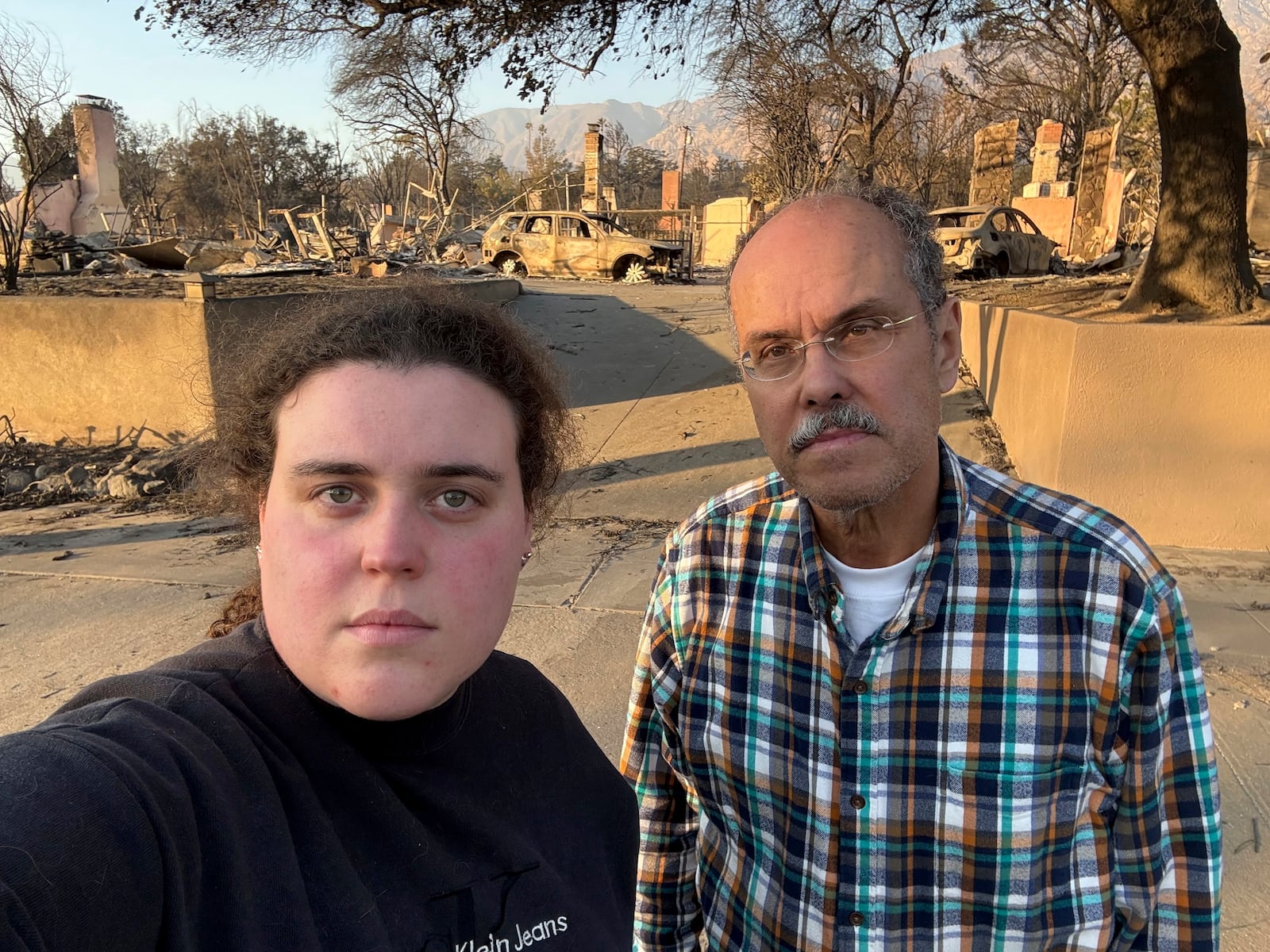 Vanessa Prata and her father, Aluizio Prata, pose for a self-portrait in Altadena, Calif., on Saturday, Jan. 11, 2025, with damage from the Eaton fire behind them. (Vanessa Prata via AP)