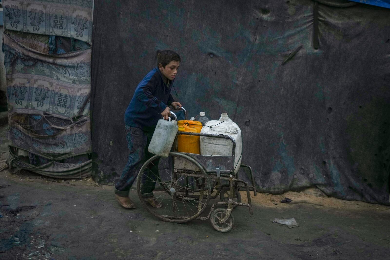 A Palestinian boy pushes a wheelchair carrying jerrycans and plastic bottles with water at a camp for displaced people in Deir al-Balah, Gaza Strip, Thursday, Dec. 12, 2024. (AP Photo/Abdel Kareem Hana)