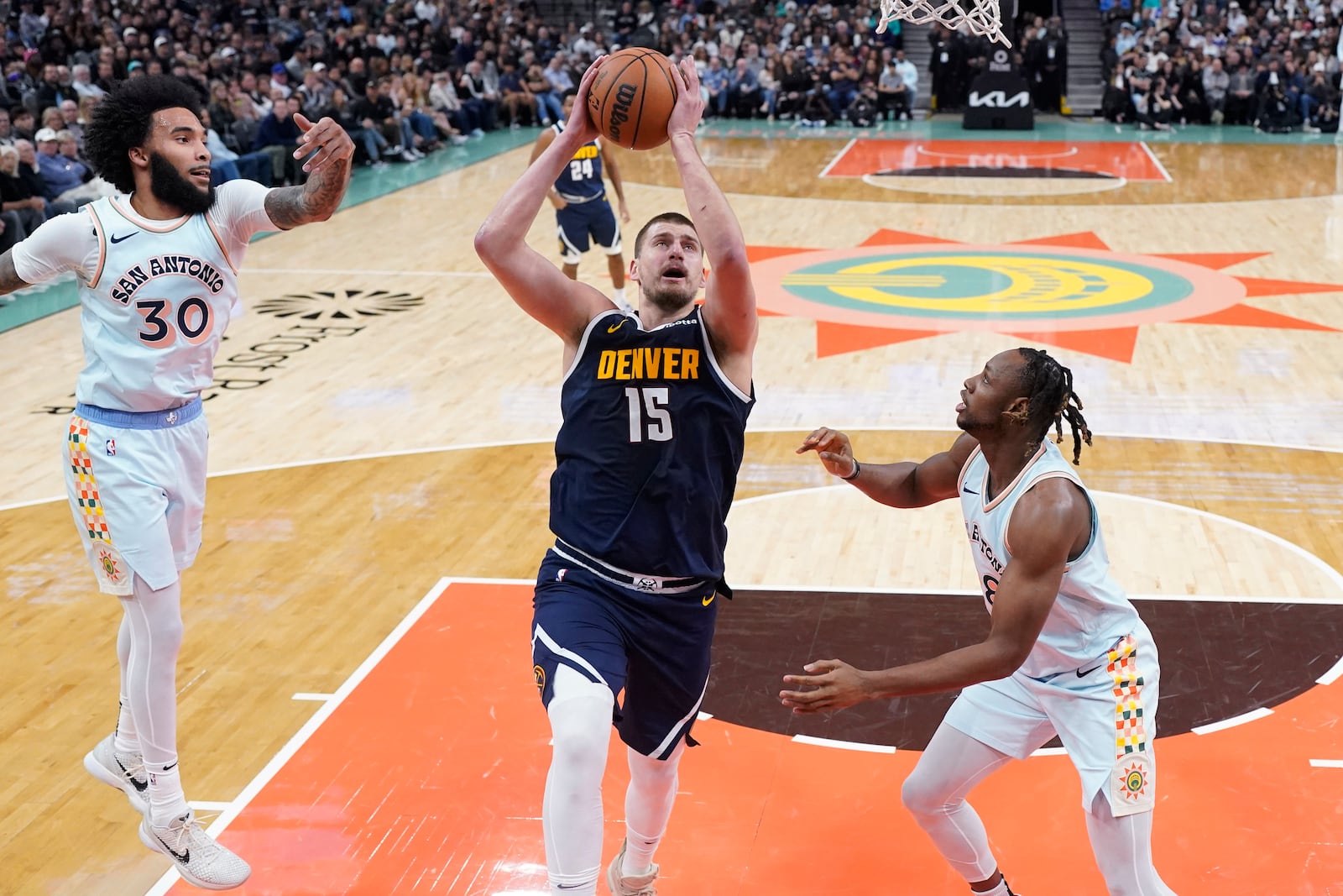 Denver Nuggets center Nikola Jokic (15) drives to the basket past San Antonio Spurs forward Julian Champagnie (30) and center Charles Bassey, right, during the first half of an NBA basketball game in San Antonio, Saturday, Jan. 4, 2025. (AP Photo/Eric Gay)