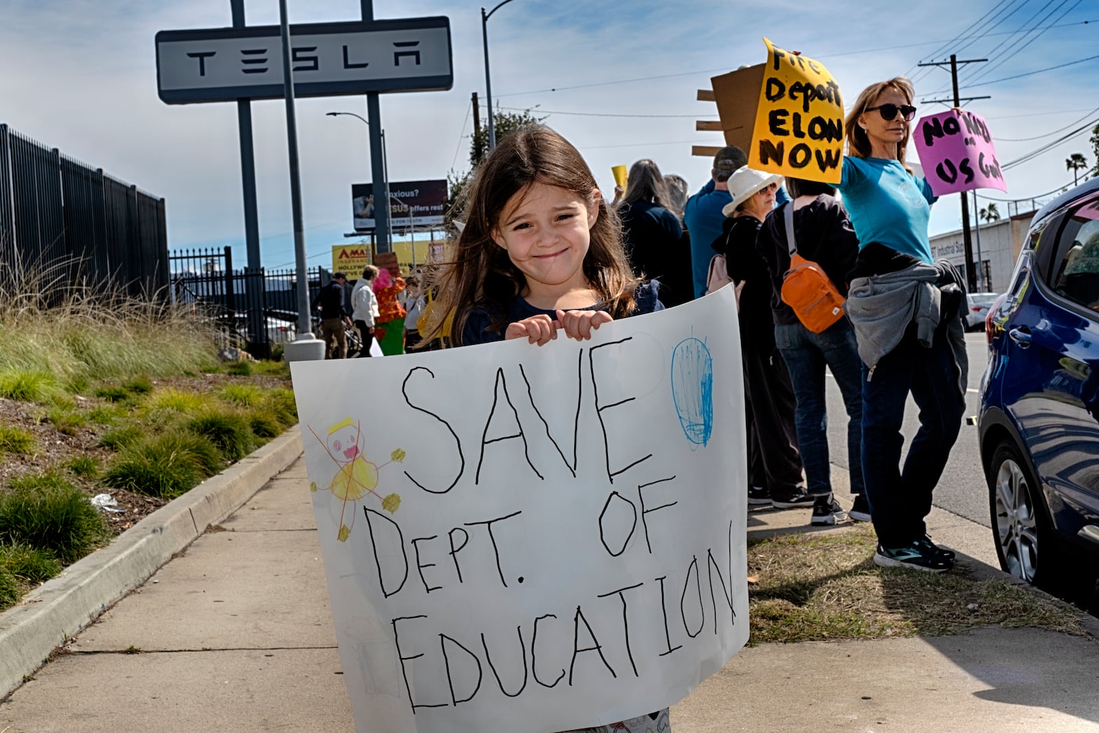 Sarah Cummings carries a protest sign to save the Department of Education while joining other protesters outside a Tesla showroom and service center in the North Hollywood section of Los Angeles on Saturday, March 15, 2025. (AP Photo/Richard Vogel)