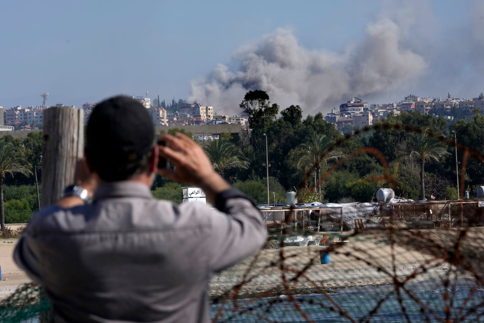 A man takes pictures by his mobile phone for the smoke rises between buildings hit in an Israeli airstrike in Burj al-Shamali village, as it seen from Tyre city, south Lebanon, Friday, Nov. 22, 2024. (AP Photo/Hussein Malla)