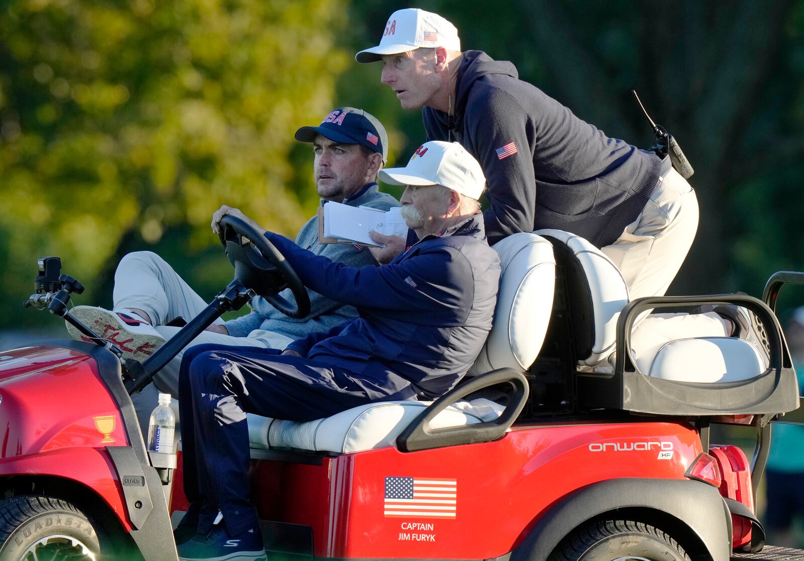 United States team captain Jim Furyk, top, grabs a ride with United States team member Keegan Bradley, left, and Mike "Fluff" Cowan driving during their fourth-round foursomes match at the Presidents Cup golf tournament at Royal Montreal Golf Club, Saturday, Sept. 28, 2024, in Montreal. (Frank Gunn/The Canadian Press via AP)