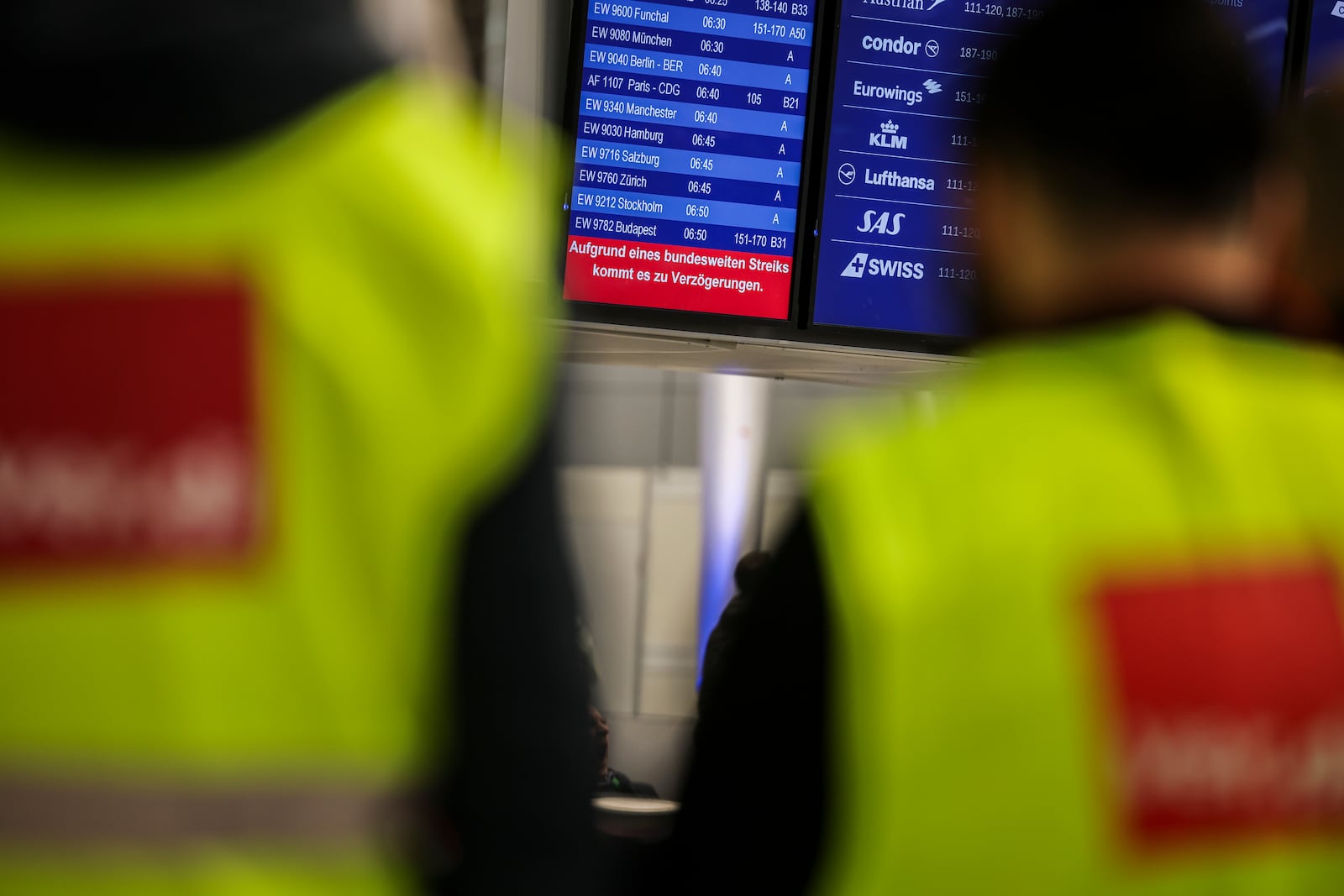 Airport employees wearing high-visibility vests from the Verdi union stand in front of a display board in the departures area at Dusseldorf Airport, Germay with a notice about the strike Monday, March 10, 2025. (Christoph Reichwein/dpa via AP)