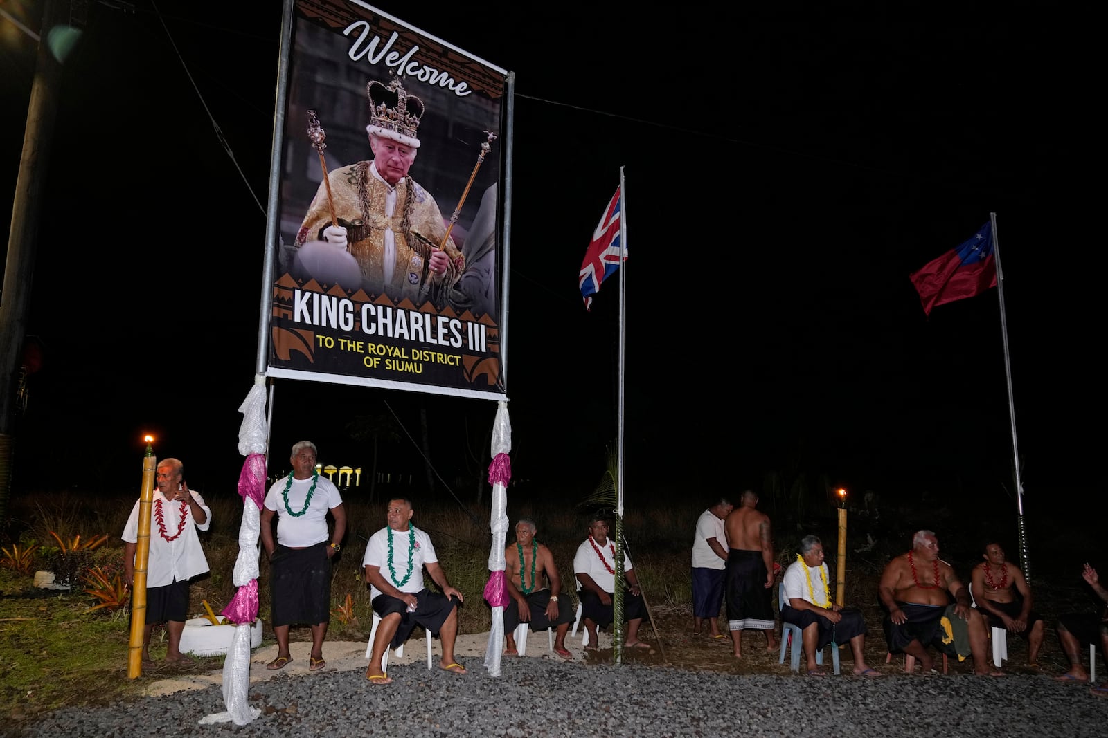 People sit under a portrait of Britain's King Charles III as they wait for his arrival and Queen Camilla in the village of Siumu, Samoa, on Wednesday, Oct. 23, 2024. (AP Photo/Rick Rycroft)