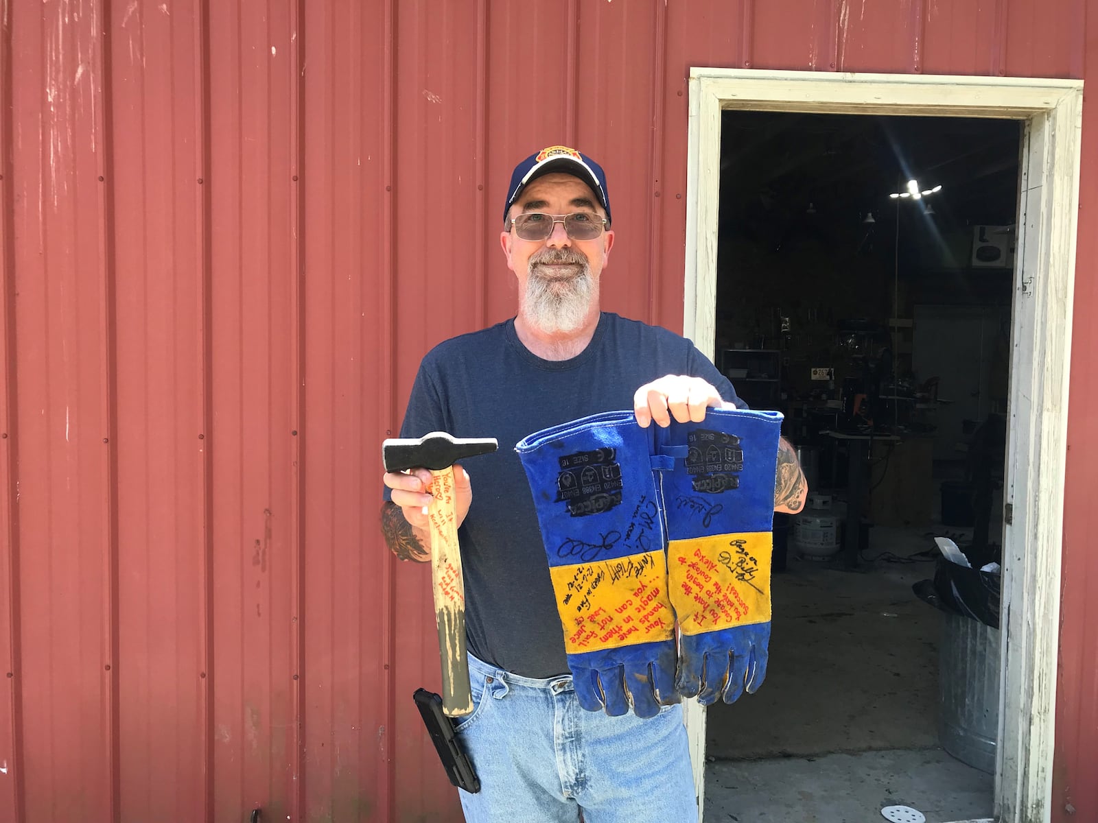 Bill Pyles stands outside of his forging workshop with the hammer and gloves he used on the show. His wife and children signed the hammer and gloves with encouraging messages.