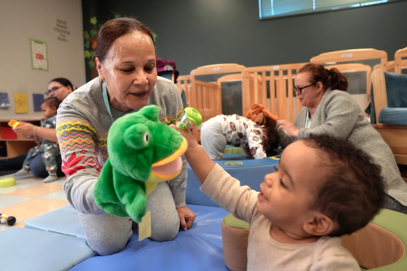 Volunteer Ydalina Luna Sosa, 62, of Providence, R.I., front left, uses a frog puppet for entertainment while supervising 10-month-old Leo Morgenweck, front right, in an early childcare program at Federal Hill House, Tuesday, Nov. 12, 2024, in Providence, R.I. (AP Photo/Steven Senne)