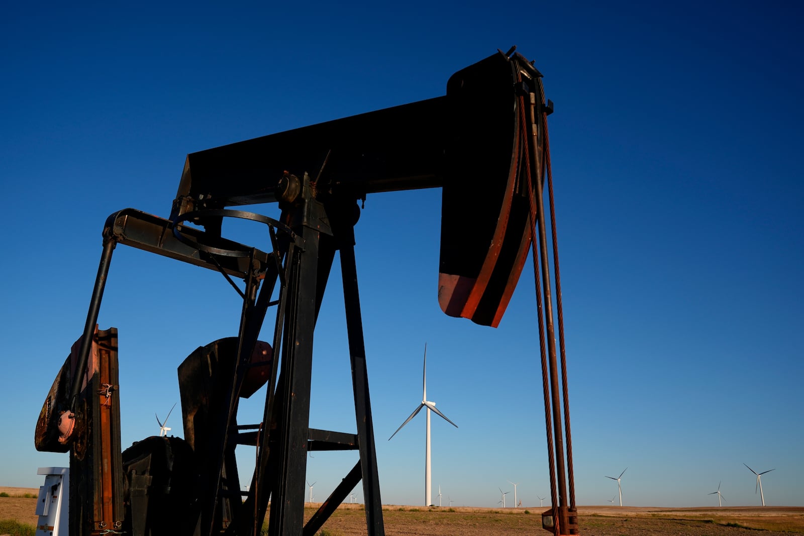 A pumpjack operates in the foreground as wind turbines at the Buckeye Wind Energy wind farm rise in the distance, Monday, Sept. 30, 2024, near Hays, Kan. (AP Photo/Charlie Riedel)