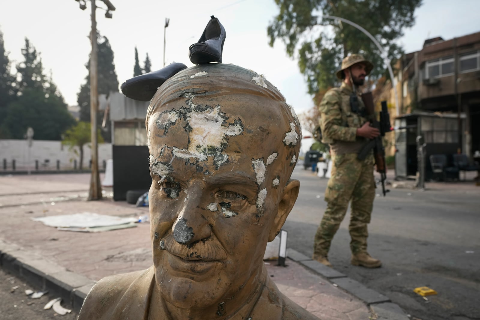 An opposition fighter in military uniform stands next to a broken bust of the late Syrian President Hafez Assad, with a pair of shoes placed on top, at a checkpoint in Damascus, Syria, Wednesday, Dec. 11, 2024. (AP Photo/Hussein Malla)