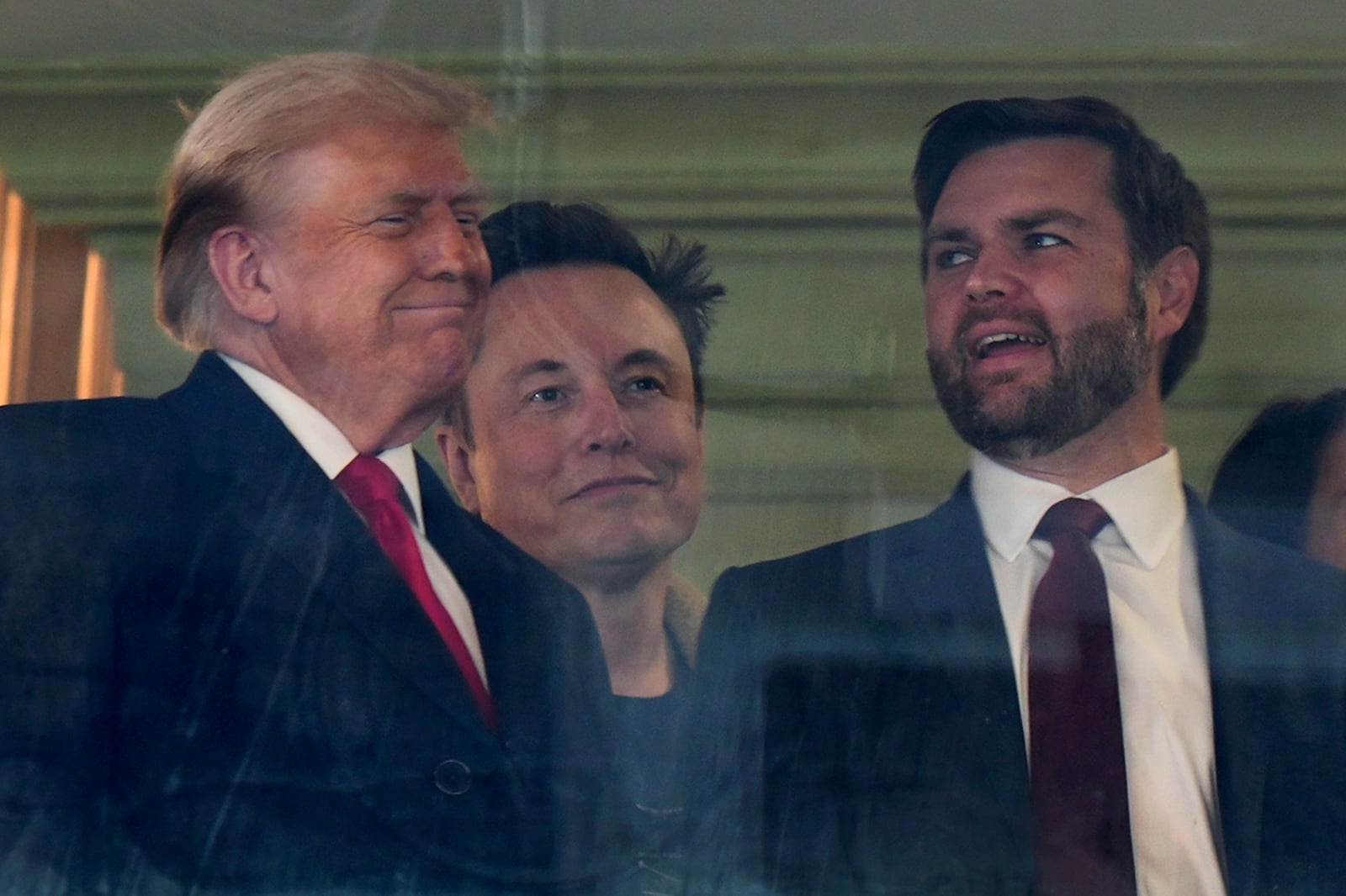 In this picture taken through a window, President-elect Donald Trump, from left, Trump's pick for the planned Department of Government Efficiency Elon Musk and Vice President-elect JD Vance attend the NCAA college football game between Army and Navy at Northwest Stadium in Landover, Md., Saturday, Dec. 14, 2024. (AP Photo/Stephanie Scarbrough)