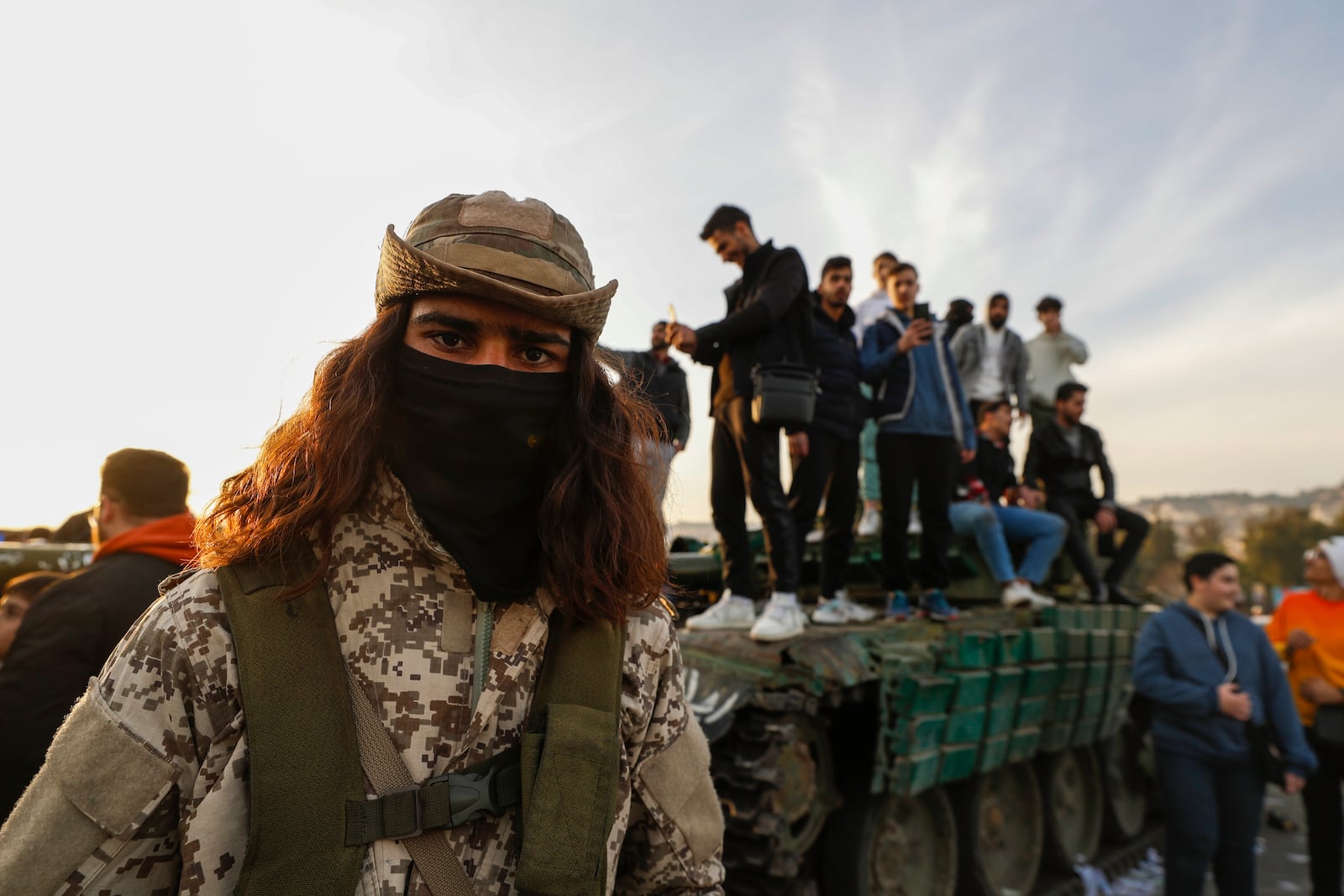 A Syrian opposition fighter, left, stands guard as Syrian citizens stand on a government forces tank, that was left on a street, during the third day of the take over of the city by the insurgents in Damascus, Syria, Tuesday, Dec. 10, 2024. (AP Photo/Omar Sanadiki)