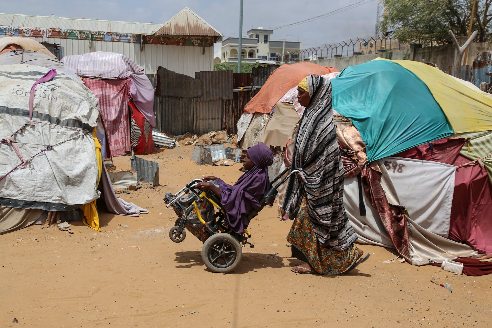 A Somali internally-displaced person (IDP) woman pushing a child in a wheelchair walks past makeshift homes in Maslah camp on the outskirts of Mogadishu, Somalia Wednesday, Feb. 5, 2025. (AP Photo/Farah Abdi Warsameh)