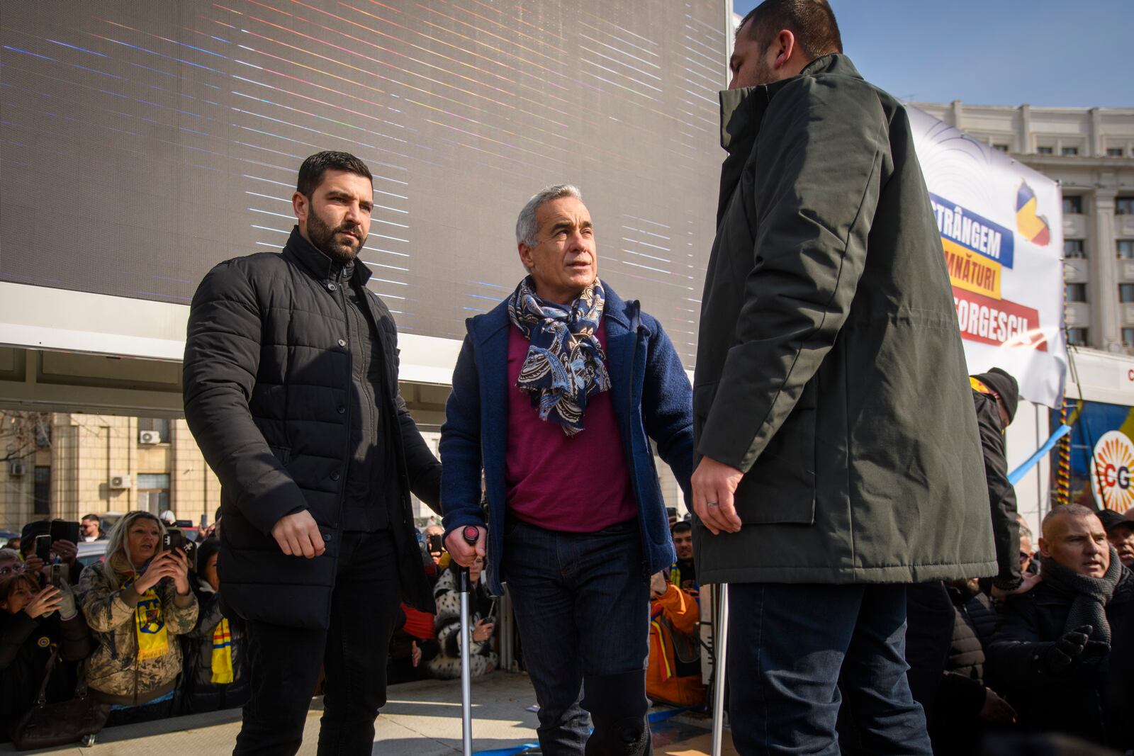 Calin Georgescu, the winner of Romania's first round of presidential election, annulled by the Constitutional Court, uses crutches while standing on a stage to greet supporters gathered for a protest outside the Romanian parliament in Bucharest, Romania, Saturday, Feb. 22, 2025. (AP Photo/Alexandru Dobre)